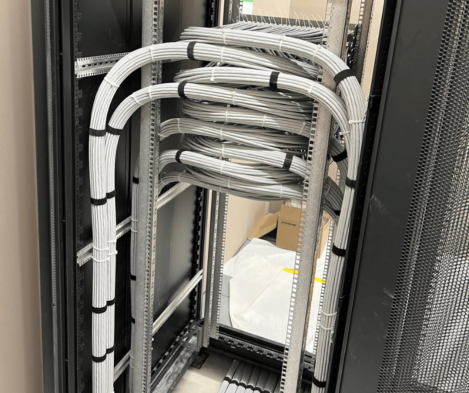 Eine Nahaufnahme zeigt das Innere eines Serverschranks oder eines Netzwerkverteilers. Im Fokus stehen zahlreiche sauber gebündelte und geführte weiße Netzwerkkabel, die in präzisen Bögen und Schleifen von oben nach unten durch vertikale Kabelkanäle verlaufen. Die Kabel sind regelmäßig mit schwarzen Kabelbindern fixiert, was eine hohe Ordnung und Struktur demonstriert. Im Hintergrund sind weitere Rack-Strukturen und ein Teil eines weiteren Schranks zu sehen, während auf dem Boden weiße Abdeckungen und ein Karton liegen, was auf Arbeiten in einem Serverraum oder Technikraum hindeutet. Das Bild betont die Sorgfalt und Präzision bei der Installation von Datenkabeln.