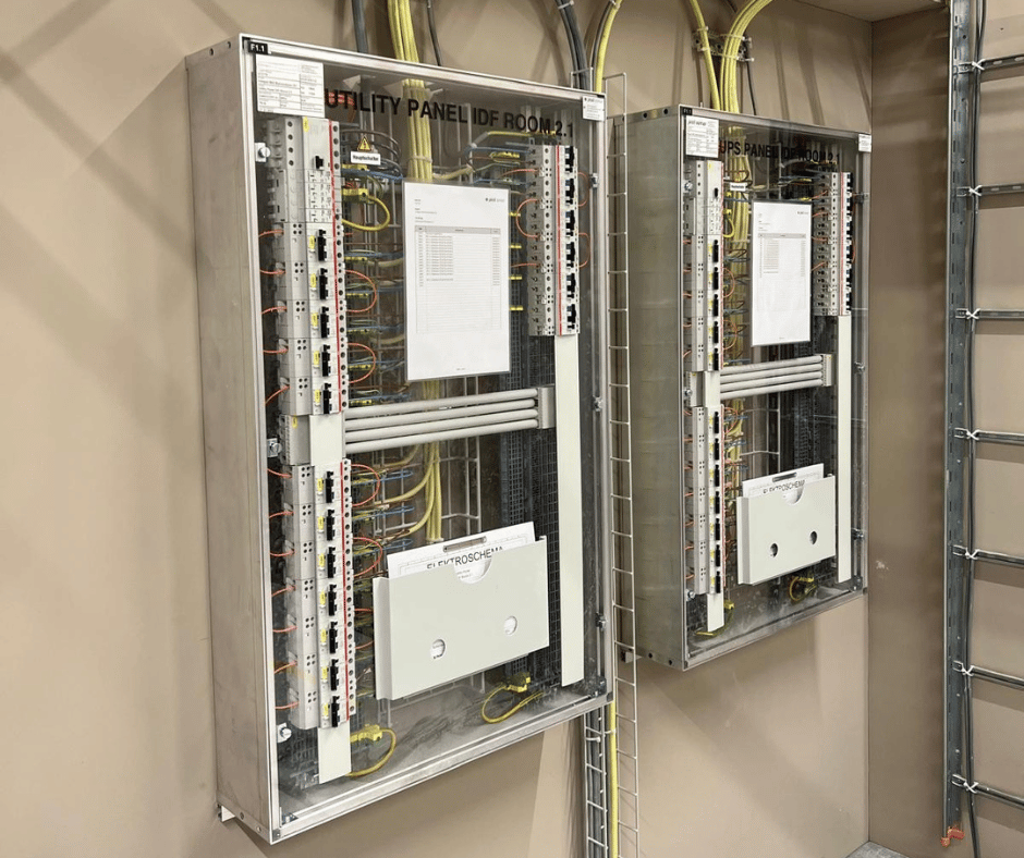 Zwei geöffnete, moderne Serverschränke oder Verteilerkästen sind in einem Technikraum nebeneinander an einer beigen Wand montiert. Der linke Schrank ist als "UTILITY PANEL IDF ROOM 2.1" und der rechte als "UPS PANEL IDF ROOM 2.1" beschriftet. Beide Schränke zeigen eine saubere, geordnete und nachvollziehbare Verkabelung mit gelben und grauen Kabeln, die an Patchfeldern und anderen Komponenten angeschlossen sind. Im Inneren beider Schränke sind Dokumente und Schaltpläne in durchsichtigen Haltern angebracht, was auf eine detaillierte Beschriftung und professionelle Organisation hinweist. Rechts im Bild ist ein Teil eines Kabelkanals oder einer Kabelpritsche zu sehen.