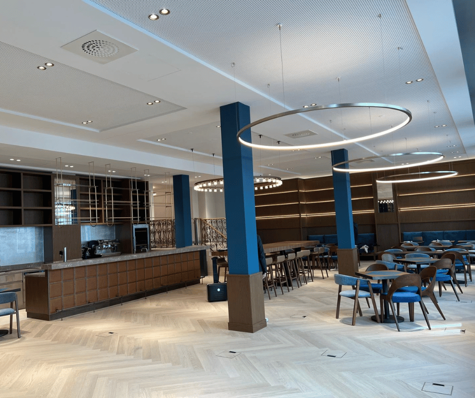 Ein weitläufiger, modern eingerichteter Innenraum, möglicherweise eine Lobby, ein Restaurant oder ein Lounge-Bereich, ist abgebildet. Der Raum ist hell beleuchtet und verfügt über eine Vielzahl von Beleuchtungselementen, darunter integrierte Deckenstrahler und auffällige, ringförmige Pendelleuchten über den Sitzbereichen. Auf der linken Seite befindet sich eine Bar oder Theke mit Kaffeemaschinen und Regalen. Der Boden ist mit einem hellen Holzparkett im Fischgrätmuster ausgelegt. Blaue Säulen und passende Sitzmöbel in Blau und Holzton verleihen dem Raum eine elegante Atmosphäre. Das Bild demonstriert die Integration von Beleuchtung, eventuell Audiotechnik (Deckenlautsprecher angedeutet) und Datenanschlüssen (Bodensteckdosen) in einer anspruchsvollen Umgebung und ist gut geeignet, um Dienstleistungen im Bereich der modernen Gebäude- und Schwachstromtechnik zu präsentieren.